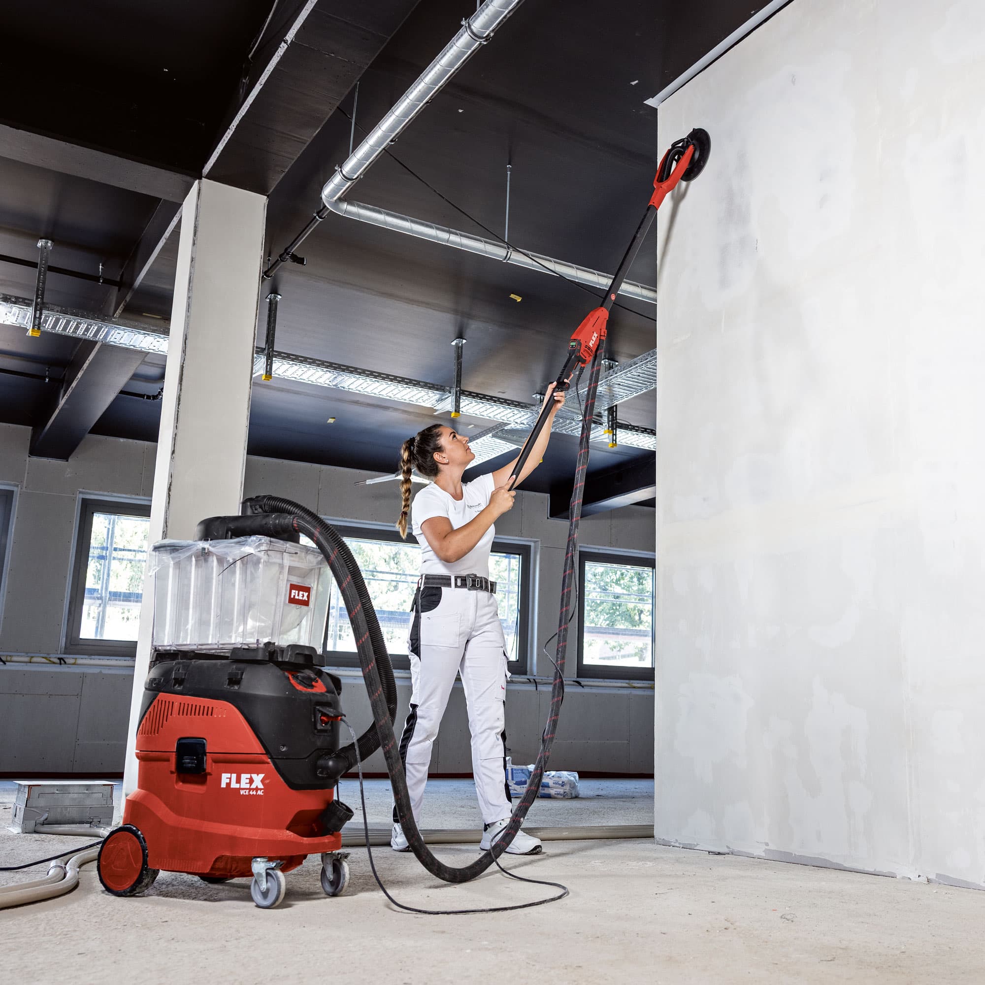 Mästermålaren Nina slipar en hög vägg med FLEX GE 6 X vägg- och takslipmaskin, ansluten till en FLEX säkerhetsdammsugare.