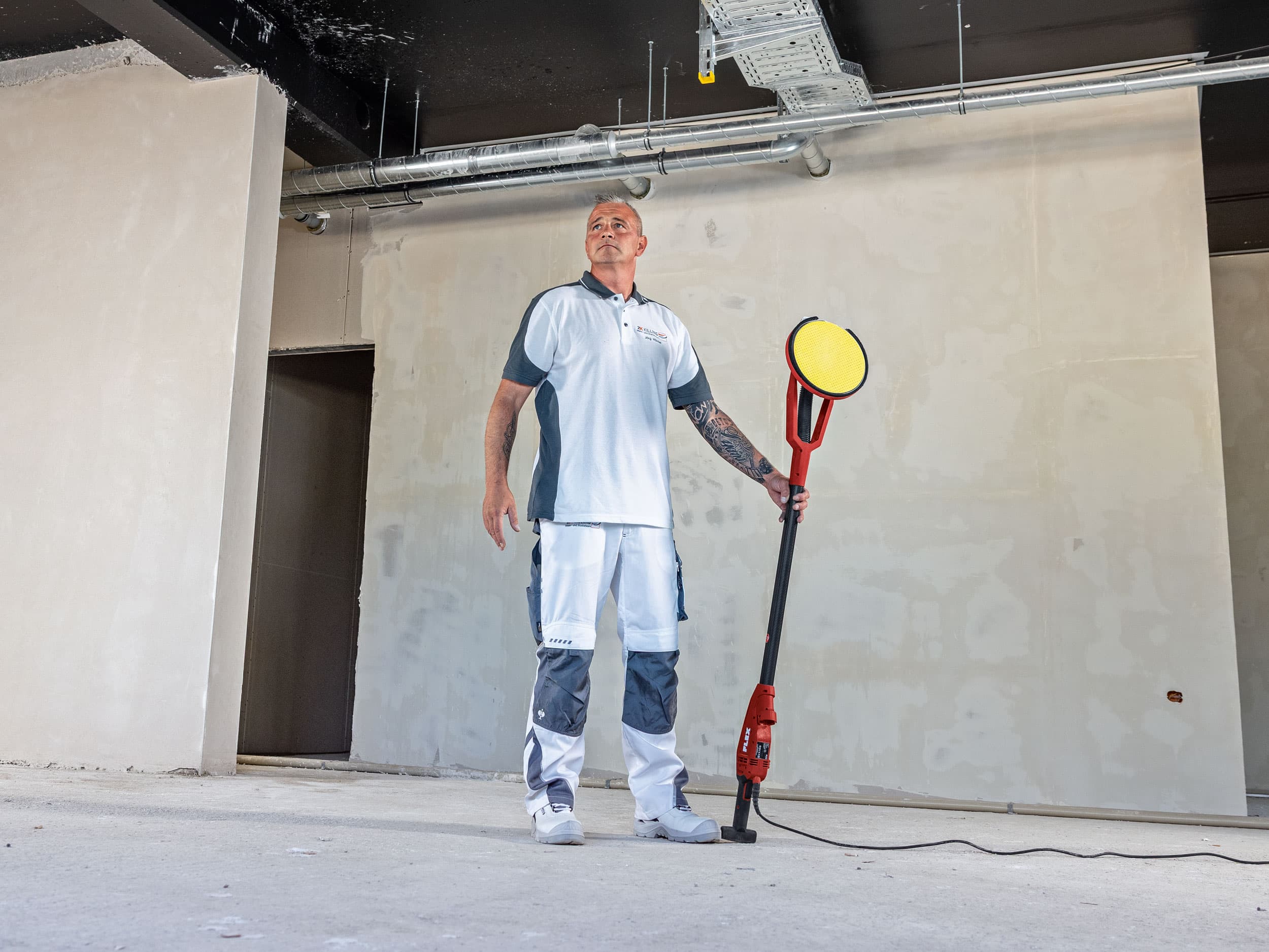 Målaren Jörg står självsäkert på byggarbetsplatsen med FLEX GE 6 R – en vägg- och takslipmaskin med roterande drivning – i handen.